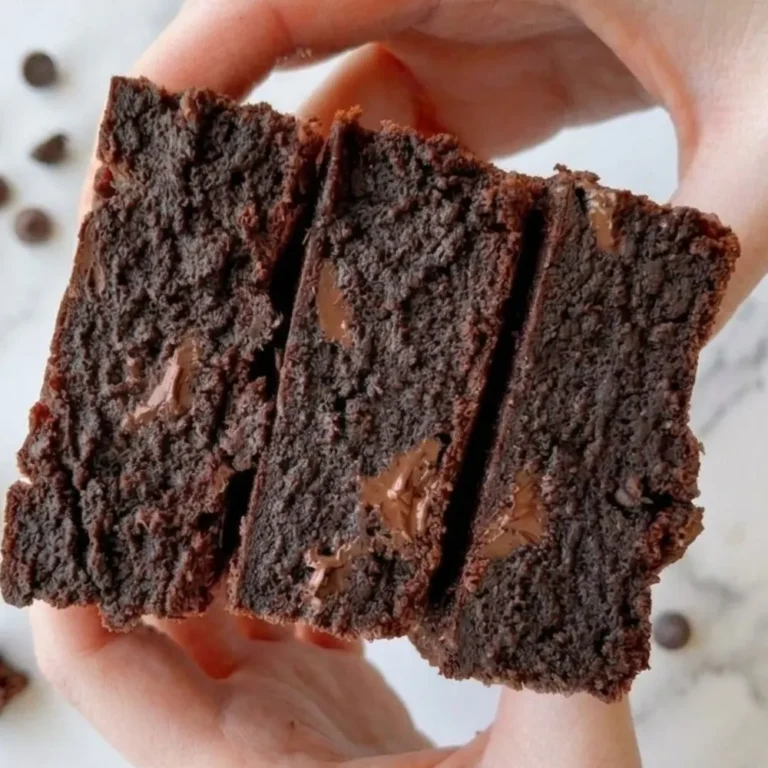 A close-up photograph of a stack of three thick, decadent homemade chocolate chip brownies, topped with flaky sea salt and chocolate shavings, resting on a dark slate board.