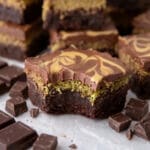A close-up view of a fudgy brownie slice with a bite taken out, showing the layers of dark chocolate, crispy green pistachio kunafa filling, and a swirled milk chocolate top.
