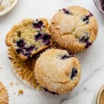 A close-up, top-down view of moist blueberry muffins on a marble counter, with one muffin cut open to reveal a fluffy interior packed with purple blueberries.
