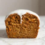 A top-down view of a single slice of carrot cake loaf on a white ceramic plate. The slice is topped with white glaze and shows small butter dots. A vintage fork is in the slice, having taken a small bite.