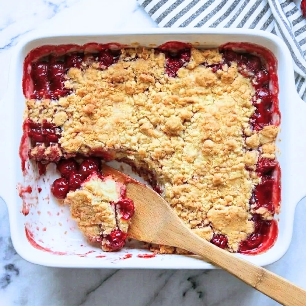 op-down view of a cherry pineapple dump cake in a white baking dish with a wooden spoon serving a portion, showing the fruit filling and golden topping.
