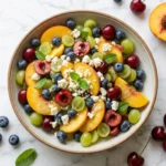 Overhead view of a vibrant Summer Fruit Salad arranged beautifully with sliced peaches, halved green grapes, dark cherries, blueberries, and crumbled feta.