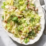A full, overhead view of the final dish from a simple apple celery salad recipe, presented on a scalloped ceramic plate with a silver fork on a marble table.